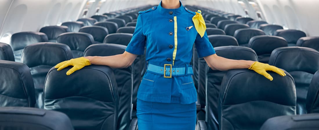 Azafata de vuelo uniformada de azul con guantes amarillos parada en el pasillo de un avión, lista para comunicarse con pasajeros internacionales.