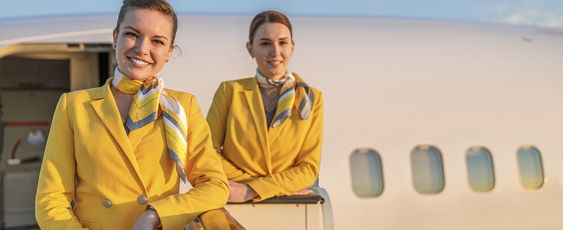 Dos TCP o azafatas sonrientes con chaquetas de uniforme amarillas y pañuelos estampados, de pie junto a la puerta de un avión.